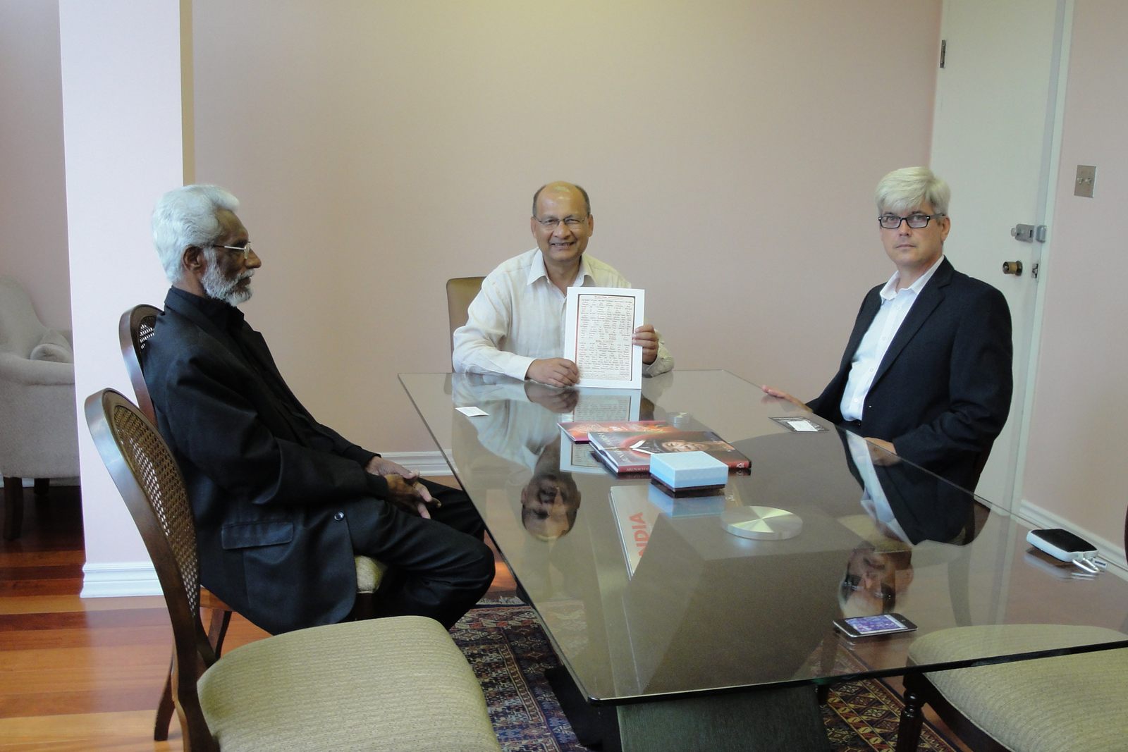 Fr. Dr. Jacob Thekeparambil, High Commissioner Vishnu Prakash and Colin S. Clarke seated around a glass table; the High Commissioner holds up a printed document.